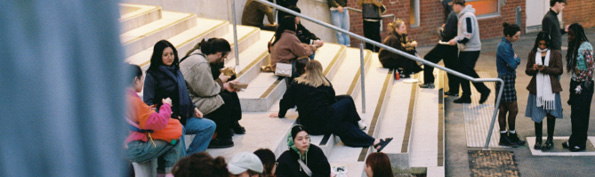 People are sitting and standing on outdoor steps and pavement near a brick building, some engaged in conversation and others using phones.

Description generated by AI