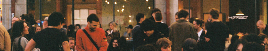 A group of people wearing casual clothing are gathered outside a building with large windows and warm string lights inside.

Description generated by AI