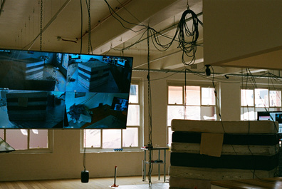 A room with large windows contains a stack of mattresses and a suspended monitor displaying multiple security camera feeds.

Description generated by AI